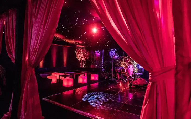 The inside of a marquee decorated for a party with a star cloth ceiling, red lighting, red curtains, and a black dancefloor