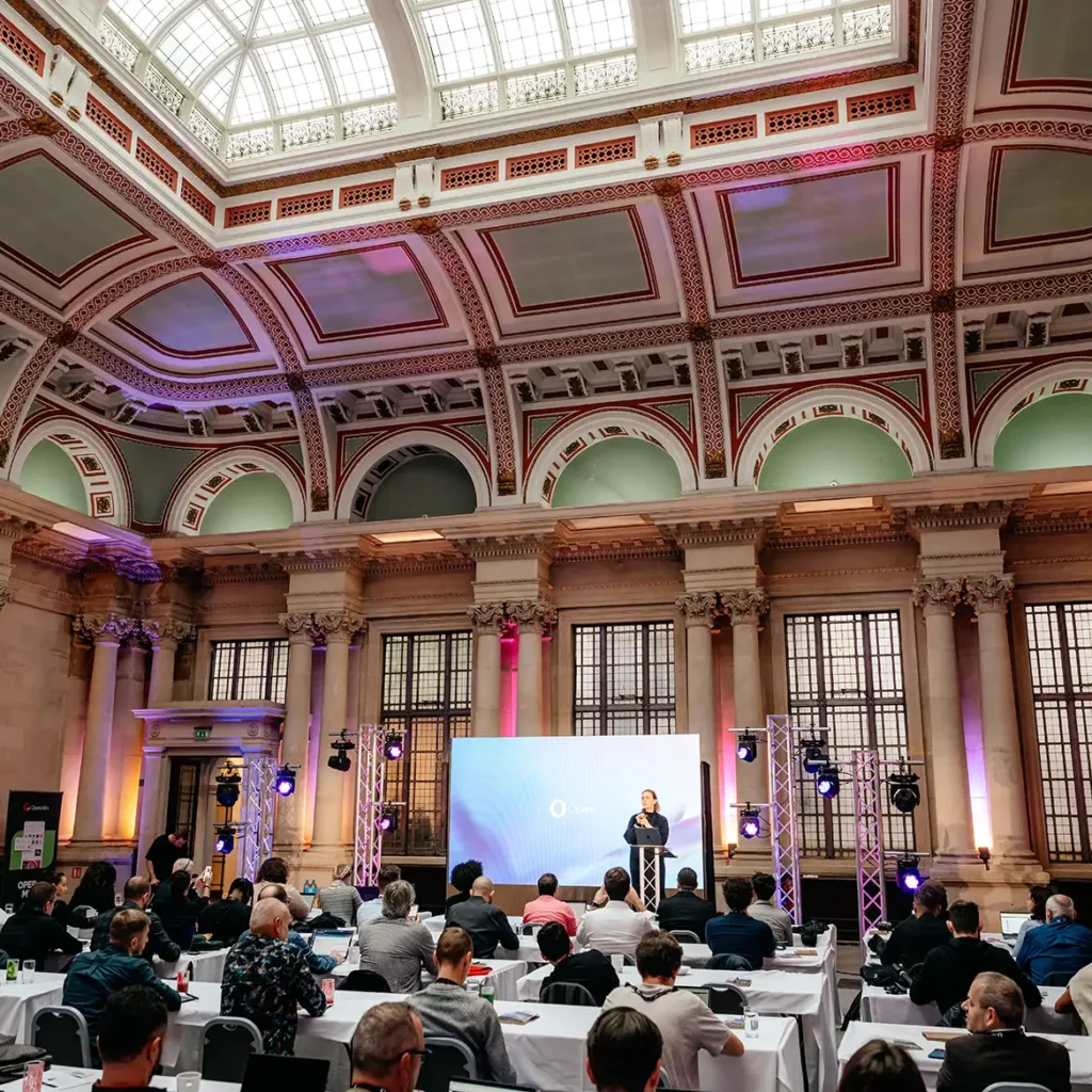Cavernous room with decorate ceiling, audience in theatre style with man presenting